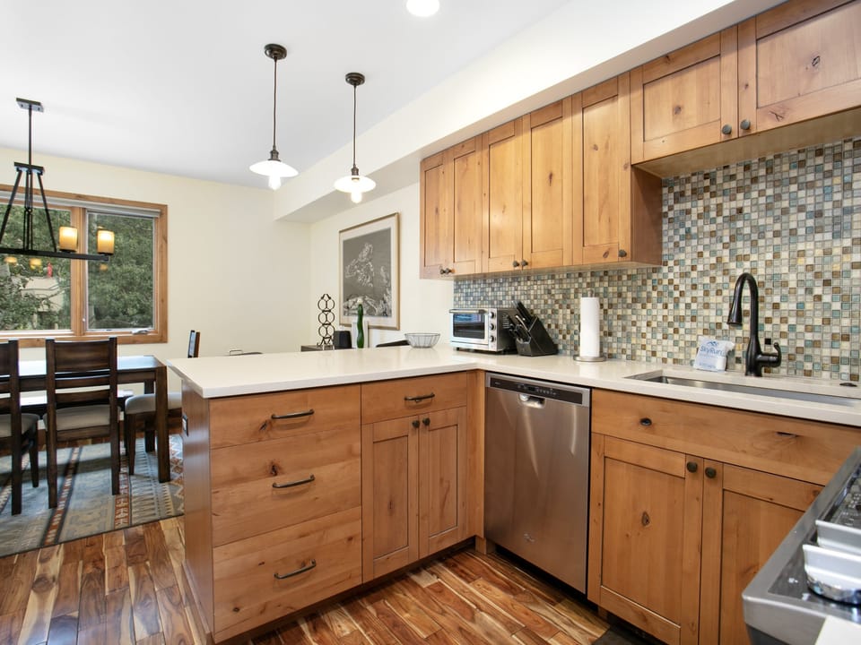 Modern kitchen with wooden cabinets, mosaic tile backsplash, and stainless steel dishwasher. Dining area with a table and chairs visible in the background. Hanging pendant lights overhead.