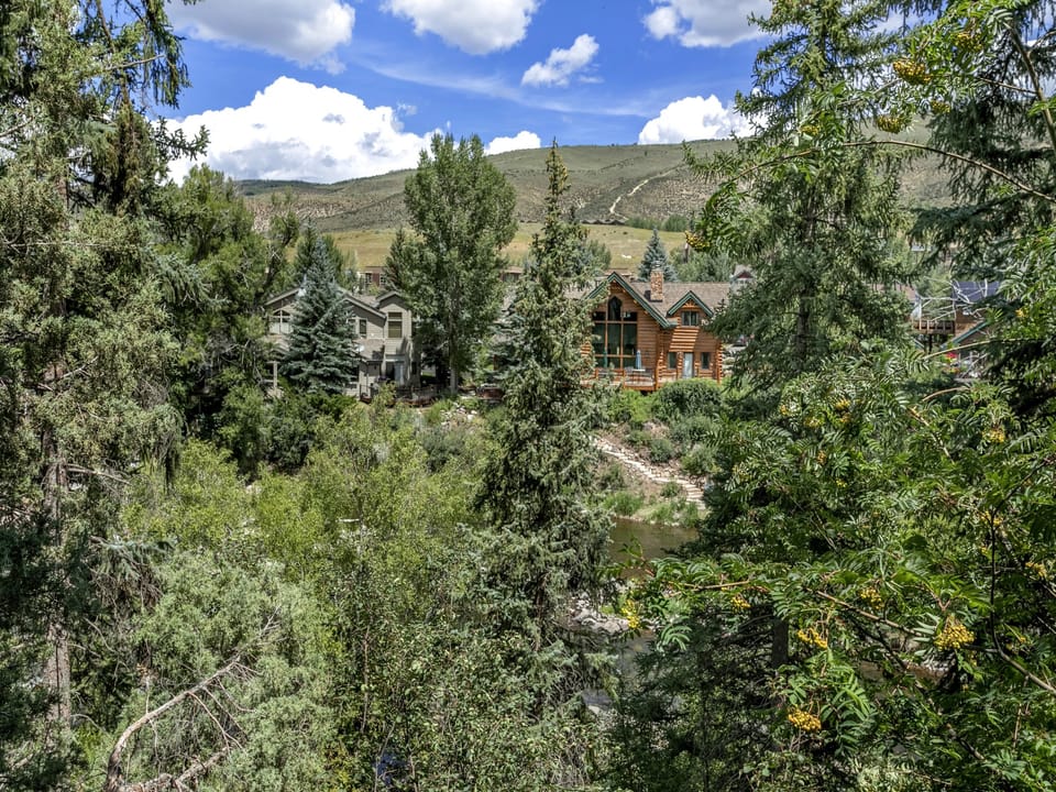 A rustic wooden house and other buildings are nestled among dense trees with a hilly background under a partly cloudy sky.