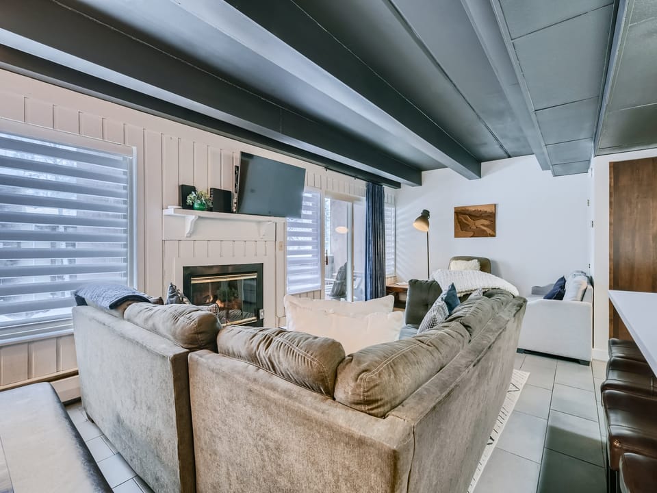 A living room with a sectional sofa, fireplace, wall-mounted TV, and a tall lamp. Large windows with blinds let in natural light.