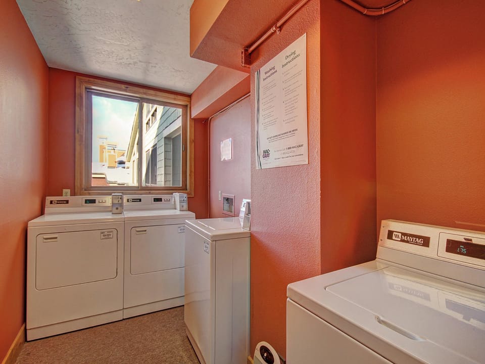 A small laundry room with orange walls, containing three washing machines, one drying machine, and a window with a view of neighboring buildings.