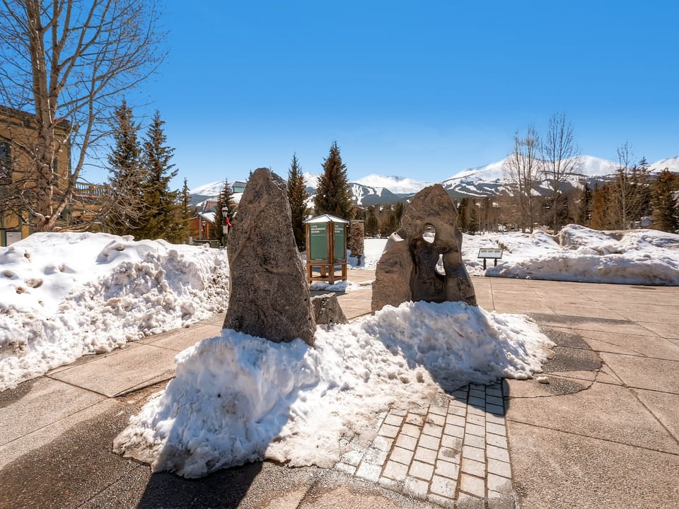 A snowy outdoor scene with large rocks standing upright on a paved area, surrounded by snow-covered paths and trees, with mountains in the background.