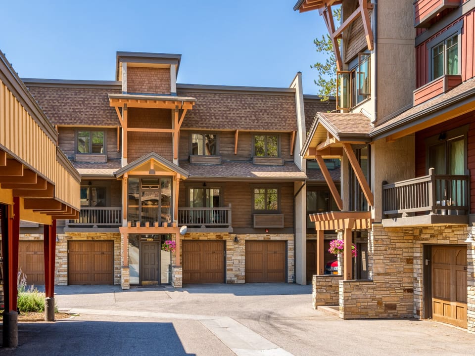 A multi-story residential building with a brown facade, balconies, garage doors, and flower pots, viewed from a paved courtyard.