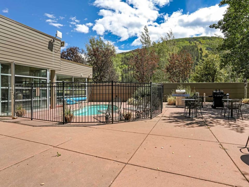 Outdoor pool area with black metal fencing, surrounded by trees and patio furniture on a paved surface. Hills and a cloudy sky are visible in the background.