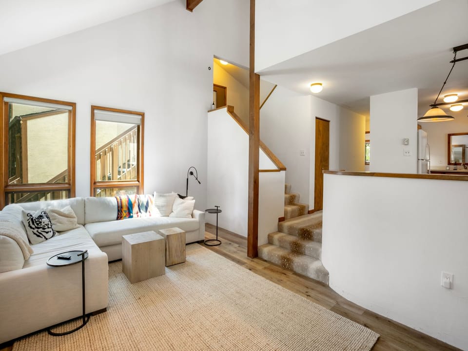 Living room with sectional sofa, wooden coffee table, and area rug. Stairs with carpet on the right lead to an upper level. White walls and multiple mirrors enhance the bright, clean space.