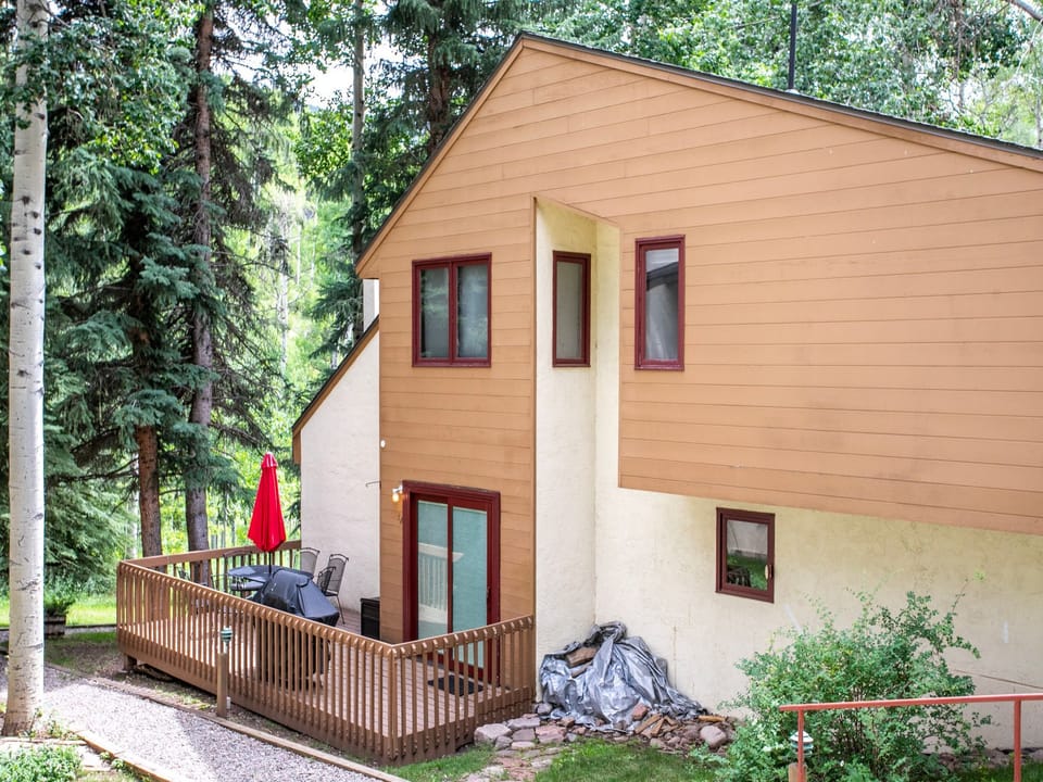 A two-story house with tan and beige siding features a wooden deck with a red umbrella, set amidst a wooded area. A pile of tarps or covers lies against the lower wall near the steps.