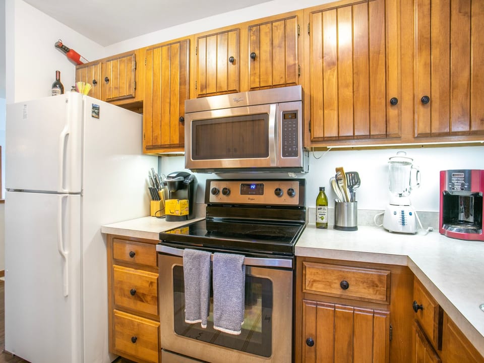 A kitchen featuring wooden cabinets, a white refrigerator, stainless steel oven and microwave, countertop appliances, and cooking utensils arranged on the countertop.