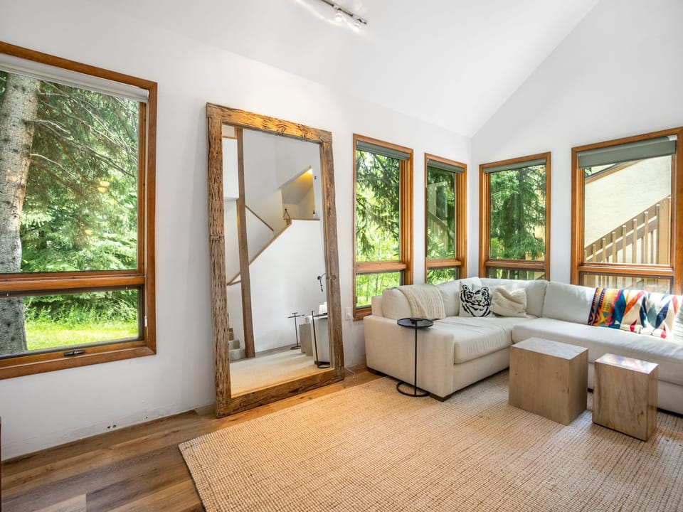 A living room with large windows, a beige sectional sofa, a wooden mirror, a rug, and a side table. The view outside the window features greenery and trees.