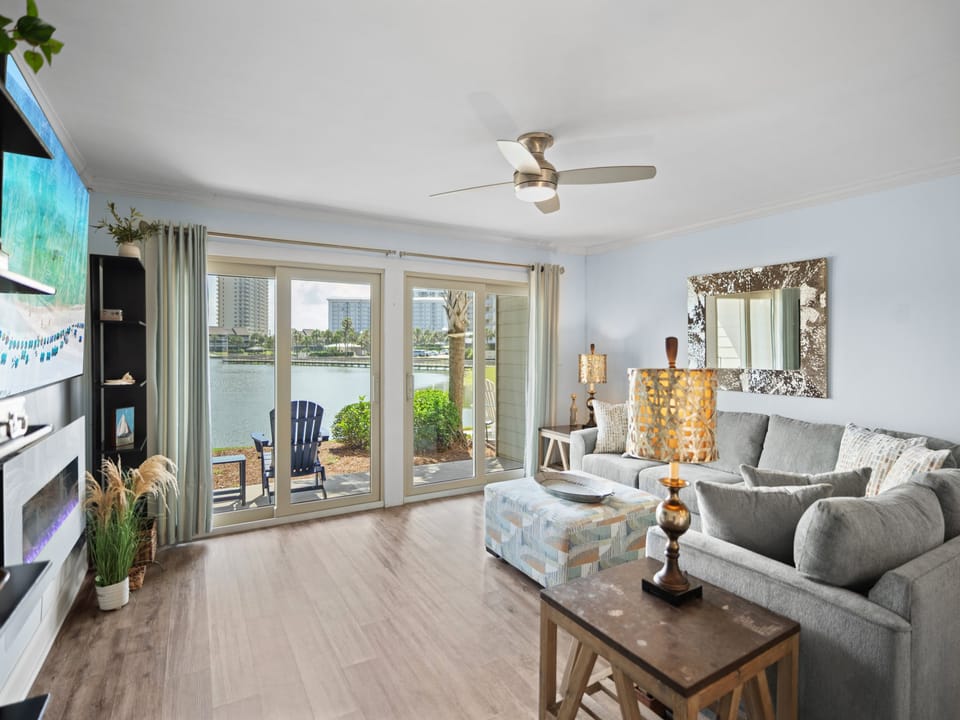 Bright, lakefront living room with large glass doors, a cozy sectional, stylish decor, and serene water views. Features patio access and warm LVP wood flooring for a relaxed, coastal feel.