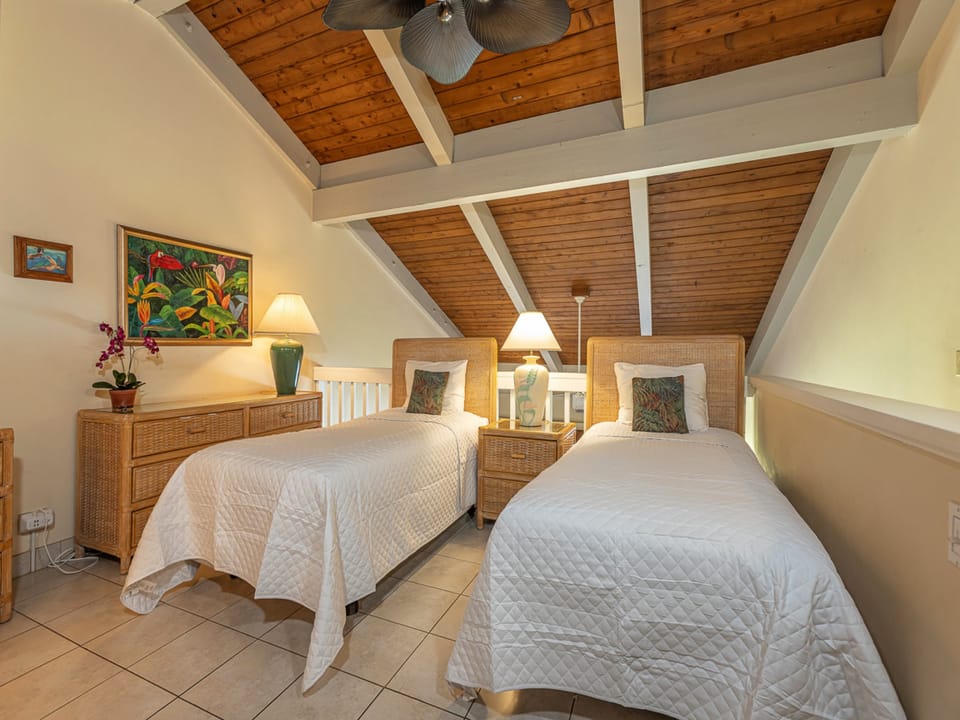 Your bright retreat features twin beds with crisp linens under a soaring wood-beamed ceiling, tropical artwork adding a splash of island charm, and wicker furnishings creating a relaxed, airy vibe.