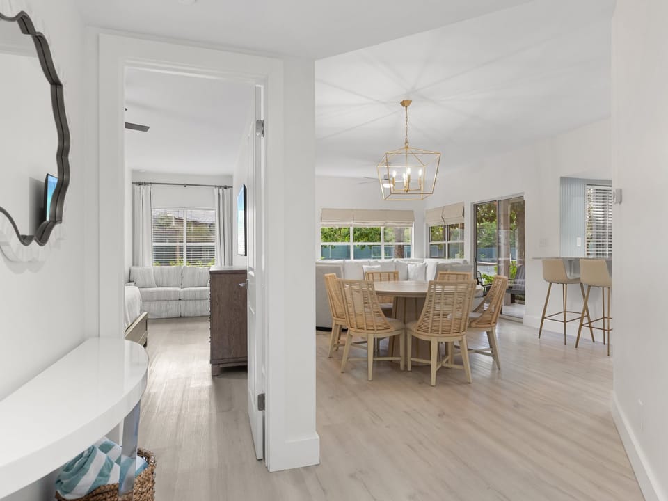 Bright and breezy, this open-concept space blends relaxed coastal style with natural light. The dining area features a round table with beachy wood tones beneath a modern gold chandelier, all flowing seamlessly into the airy living room beyond.