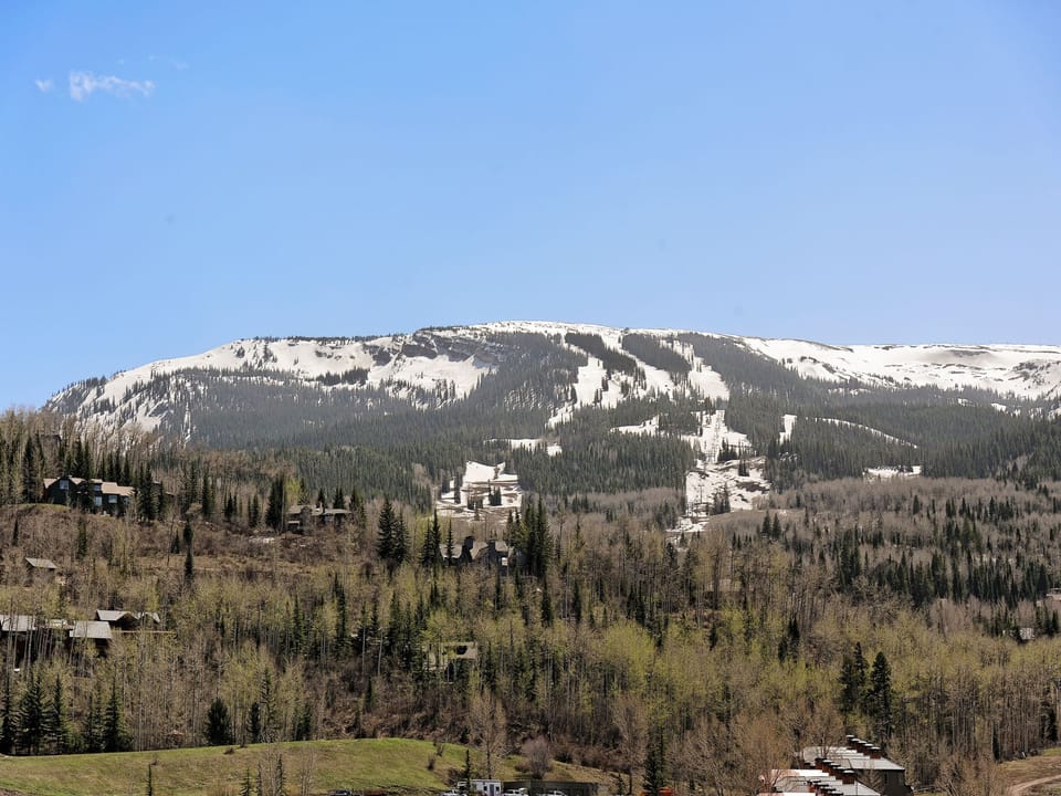 A mountain landscape showing a snow-covered ridge, partially forested slopes, and scattered buildings nestled among trees under a clear blue sky.