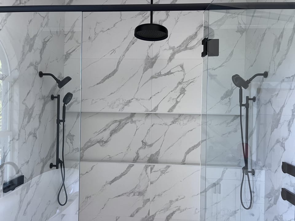 A modern bathroom features a glass-enclosed shower with white and gray marble walls, dual black showerheads, and sleek black fixtures.