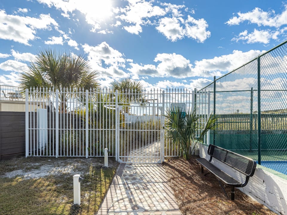 Poolside seating area with tropical palms and chain-link fencing under a bright blue sky with scattered clouds.