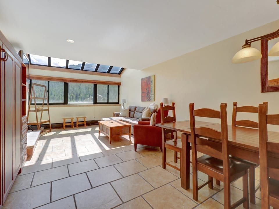 Spacious open-concept living and dining area flooded with natural light from a full-length skylight. Comfortable seating and rustic wood furnishings create an inviting mountain retreat.