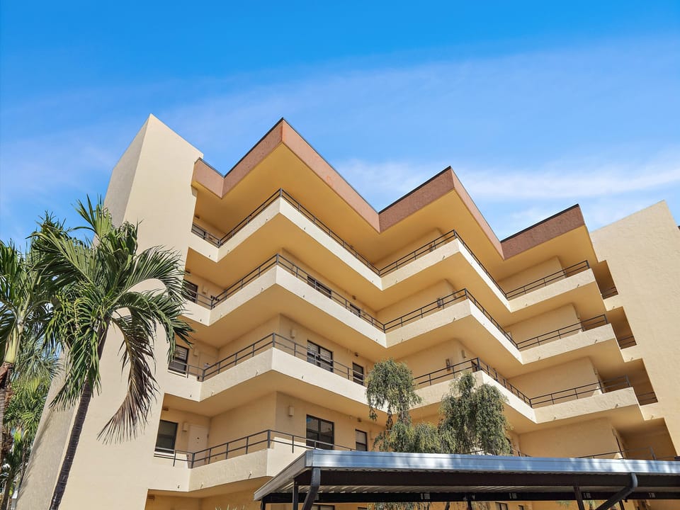 Multi-story vacation rental building with terraced balconies and tropical landscaping under a clear blue sky.