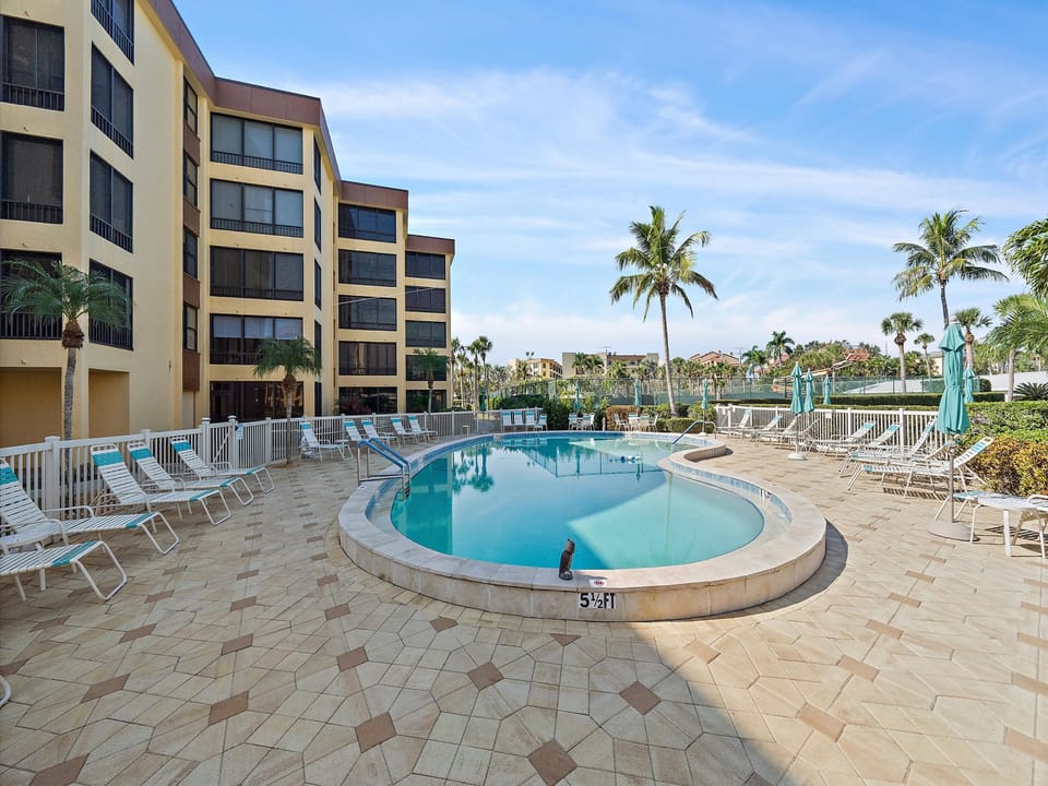 Tropical resort grounds featuring a sparkling swimming pool surrounded by palm trees and modern buildings in a sunny Florida setting.