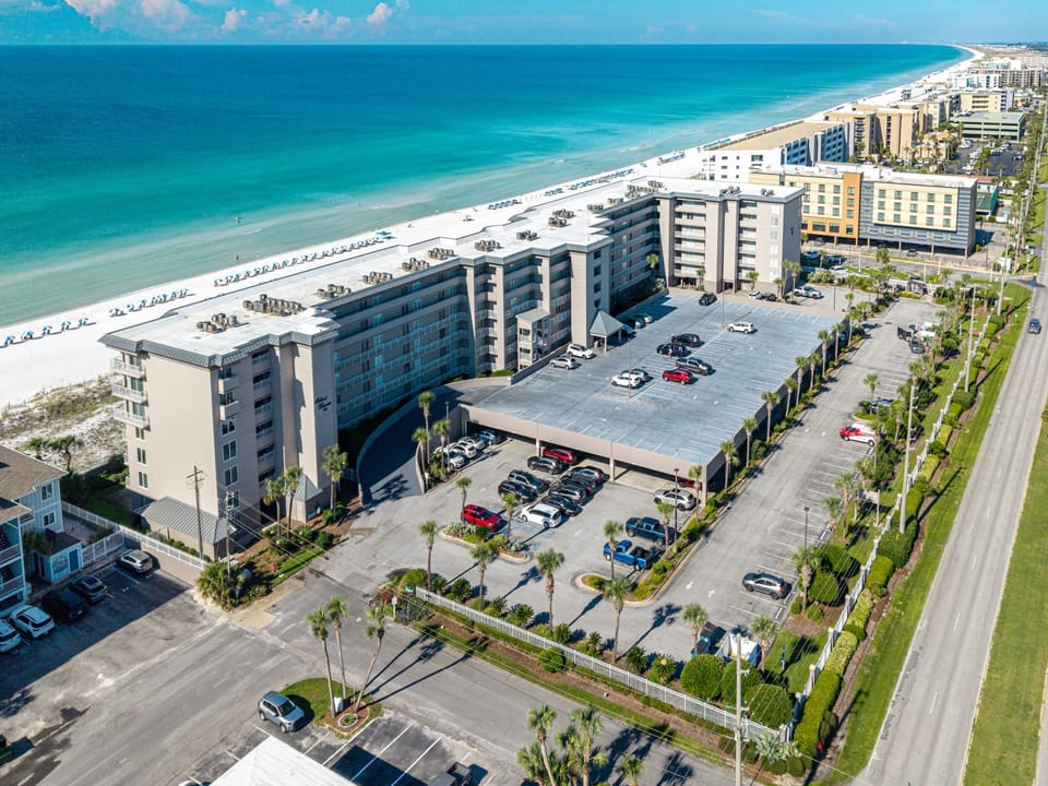 An aerial perspective reveals the property's beachfront location along white sand shores and turquoise waters, with convenient parking and nearby coastal amenities stretching along the coast.
