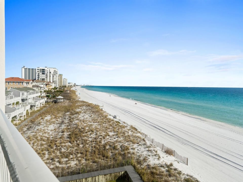 Pristine white sand beach stretches along turquoise waters with coastal buildings and natural dune vegetation.
