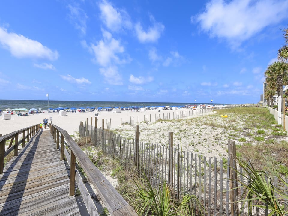 Step onto the wooden walkway leading to the sandy beach, where colorful umbrellas and beach chairs await your relaxation