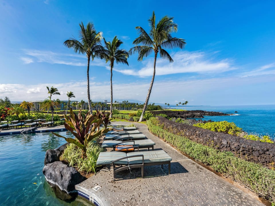 Your private oceanfront pool deck awaits with lounge chairs perfectly positioned beneath swaying palms, where turquoise waters meet black lava rock shores under clear Hawaiian skies.