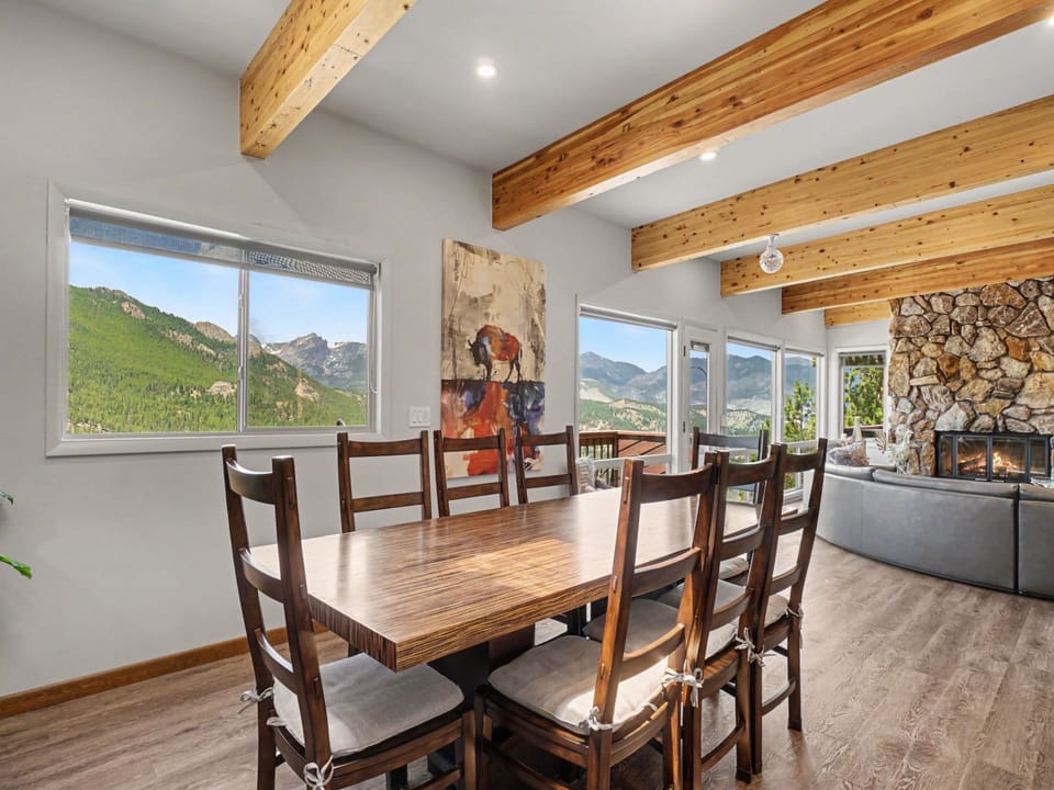 Gather around a beautiful wood table beneath exposed beams, where expansive windows frame breathtaking mountain views and a stone fireplace adds warmth to your dining experience.