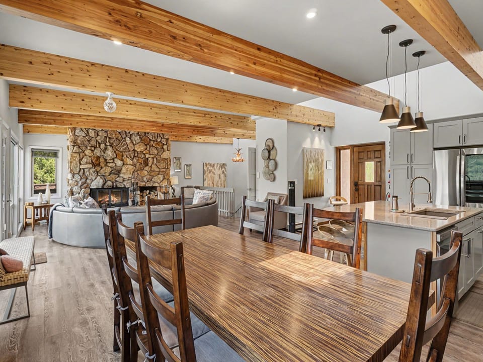 Warm wood beams stretch overhead, framing your open-concept gathering space where rustic stone fireplace meets modern kitchen—perfect for making memories around the handcrafted dining table.