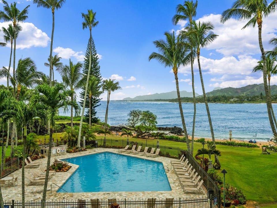 A stunning oceanfront property featuring a pristine pool surrounded by swaying palms, with dramatic mountain views across turquoise waters creating an idyllic tropical paradise setting.