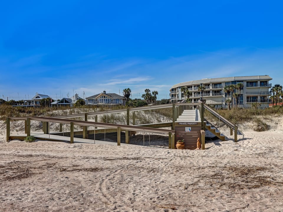 A wooden boardwalk leads from the sandy beach through natural dunes to the beachfront property, with palm trees and coastal buildings visible beyond.