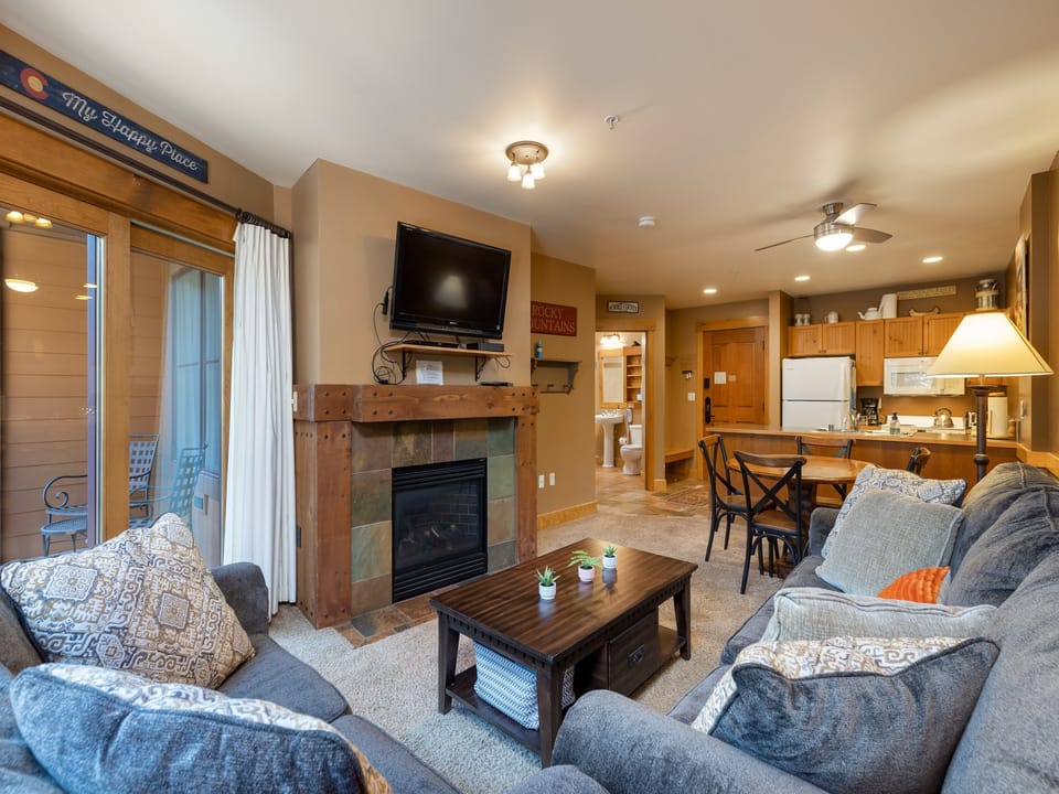 A cozy living room with grey sofas, a wooden coffee table, a fireplace, and a wall-mounted TV. The room leads into an open kitchen with wooden cabinets and a dining area with a table and chairs.