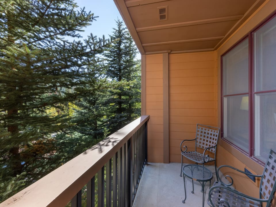 A second-floor balcony with a metal table and chairs overlooks a forested area with tall evergreen trees. The building has wooden siding and a railing.