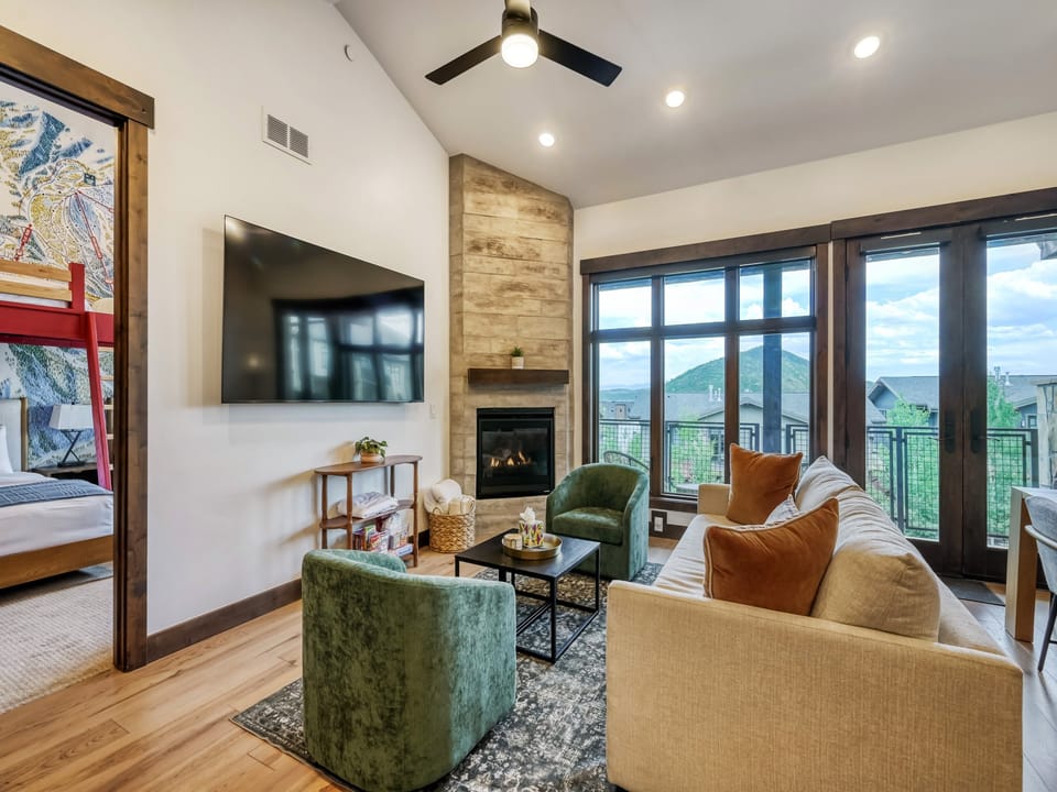 Relax in your bright living room with vaulted ceilings, a cozy fireplace, modern seating, and stunning mountain views framed by expansive windows and balcony access.