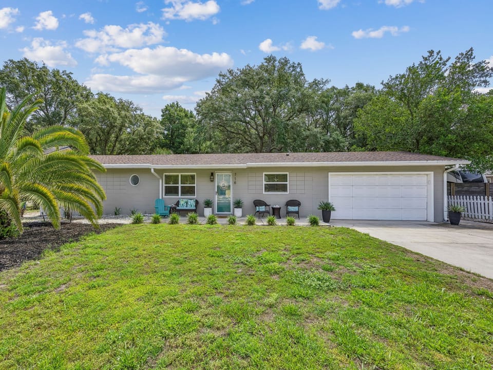 A charming single-story home with inviting front porch seating, lush green lawn, and mature trees providing natural shade in a peaceful neighborhood setting.