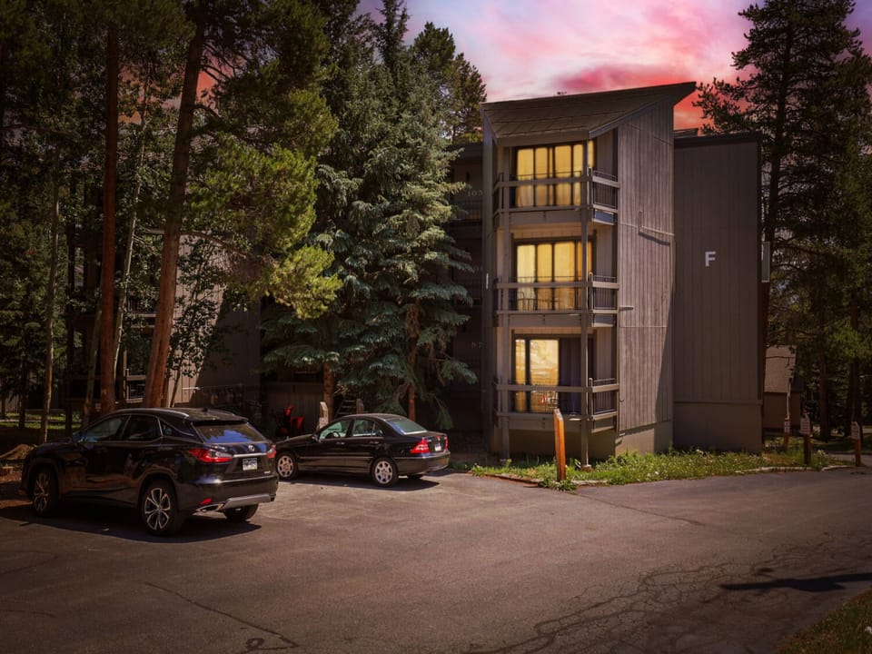 Modern mountain retreat nestled among towering pines, illuminated against a stunning sunset sky, with convenient parking steps from the entrance.