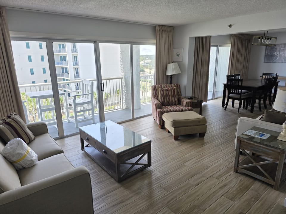Relax in this bright, open living space where comfortable seating invites you to unwind, and your private balcony overlooks coastal greenery and nearby buildings—perfect for morning coffee or evening breezes.