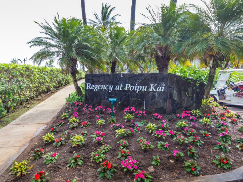 The welcoming entrance sign greets visitors to Regency at Poipu Kai, set against lush tropical landscaping with vibrant flowers.