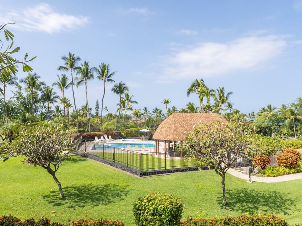 A lush tropical oasis with swaying palms surrounding the sparkling pool and thatched pavilion, framed by vibrant green lawns and colorful island landscaping.