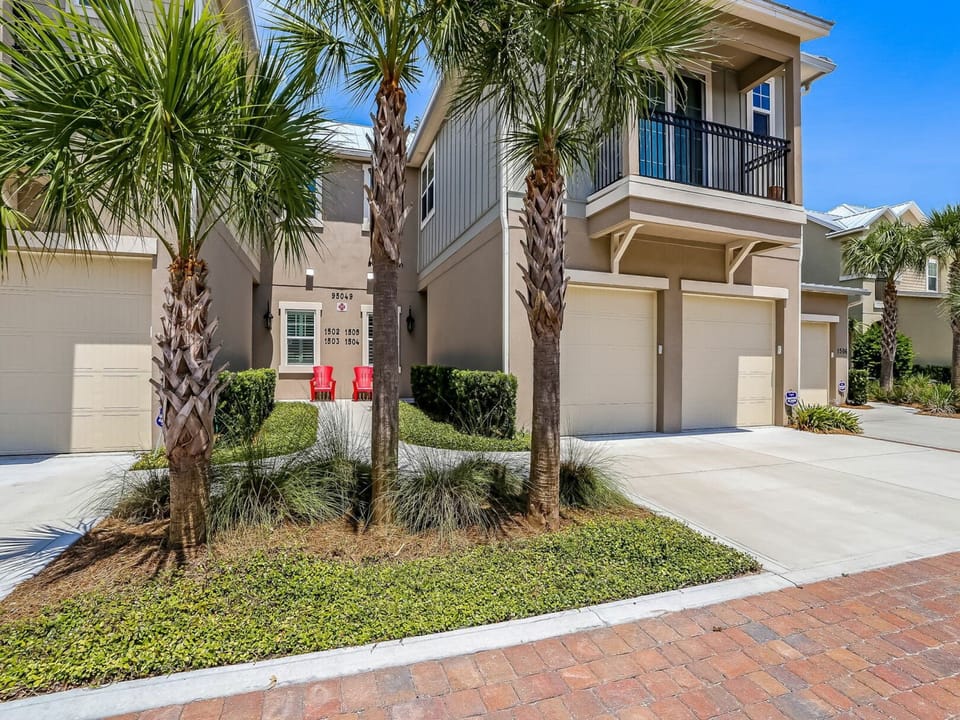A two-story vacation townhome with a private garage and balcony, surrounded by palm trees and coastal landscaping in a peaceful residential community.