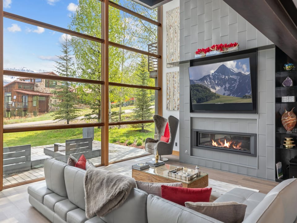 This living room features soaring windows framing neighboring properties and natural surroundings, with modern seating around a sleek fireplace creating an inviting space to gather and unwind.