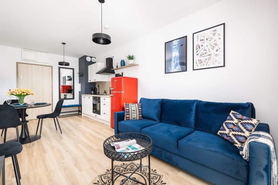 A bright living room featuring a blue sofa, a red fridge in the kitchen area, and a minimalist dining table.
