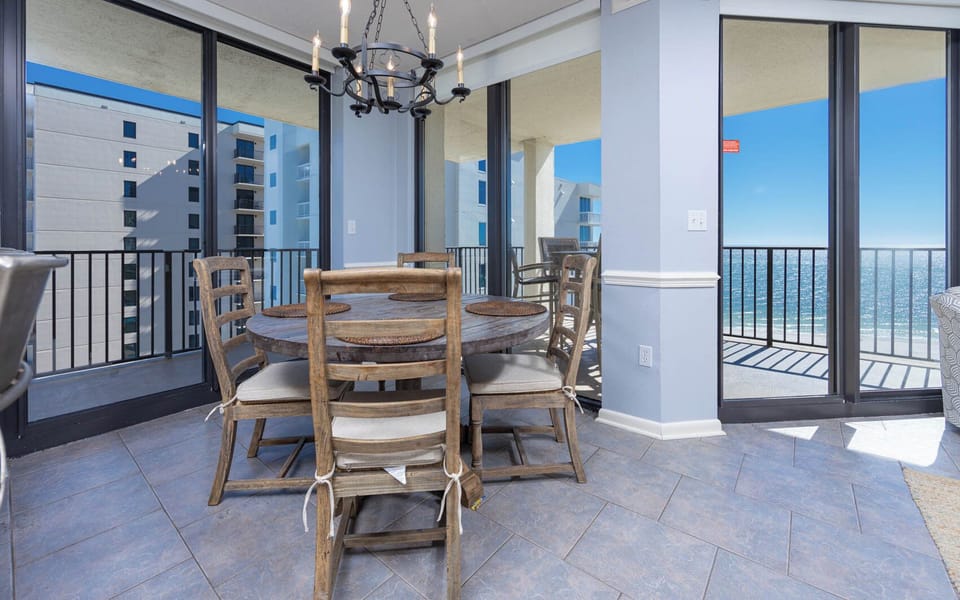 Dining area features a round wooden table with rustic chairs, offering a view of the beach through expansive glass doors
