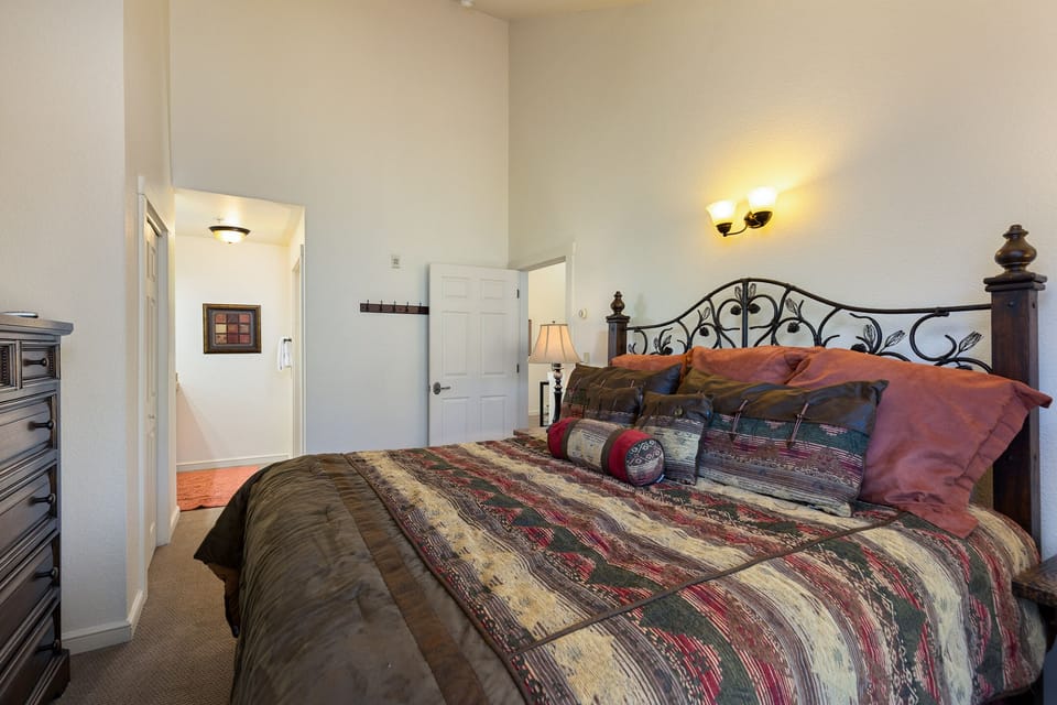 A bedroom with a patterned bedspread, decorative pillows, a wrought iron headboard, a dresser on the left, a lit lamp on the right, and a hallway leading to another room.