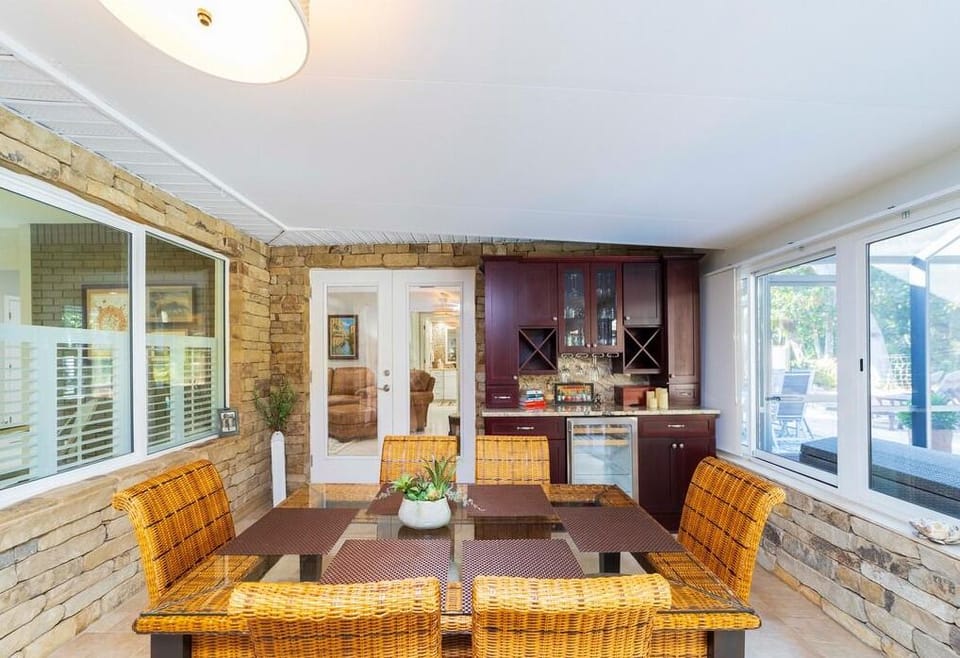 Glass-topped dining table surrounded by woven rattan chairs in a stone-walled sunroom with built-in cabinetry.