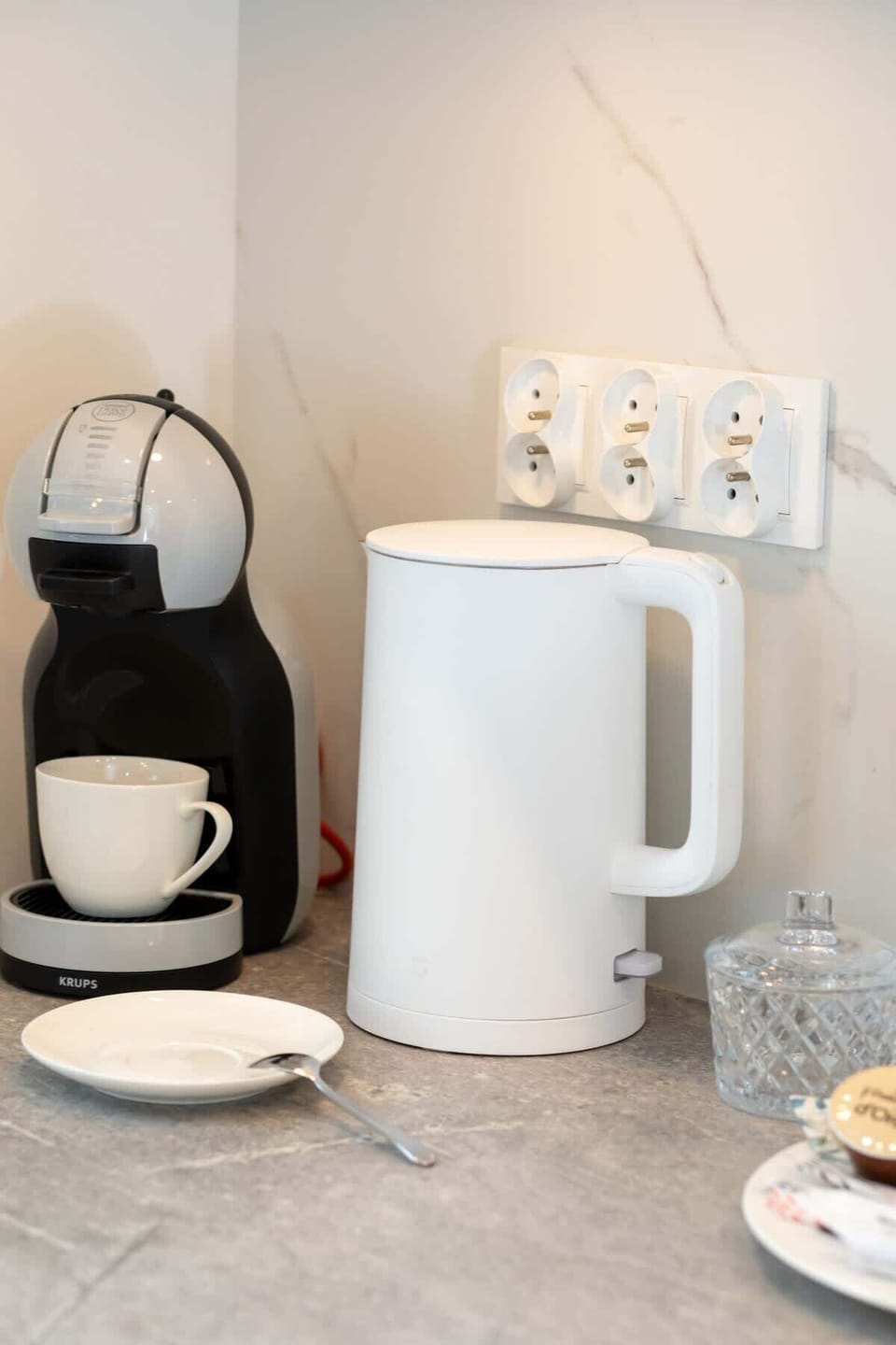 A compact coffee station with a white kettle, cups, and a selection of tea and coffee.

