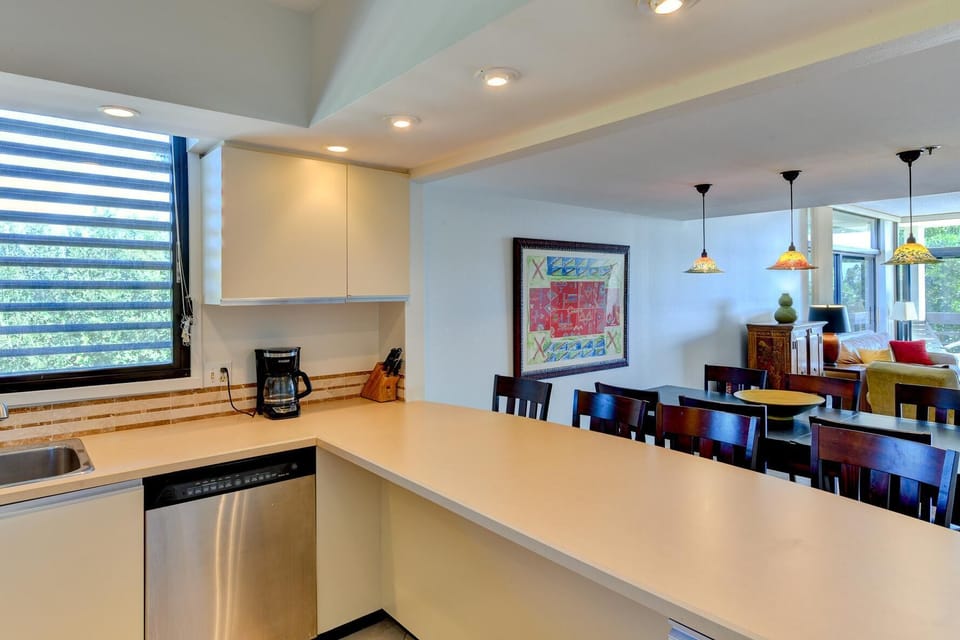 Kitchen counter with light beige laminate surfaces and stainless steel dishwasher under a window with layered blinds.