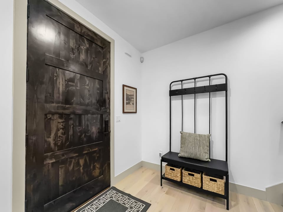 A minimalist entryway features a dark wooden door, a black metal coat rack with a cushion, and three woven storage baskets, alongside a framed picture on a white wall.