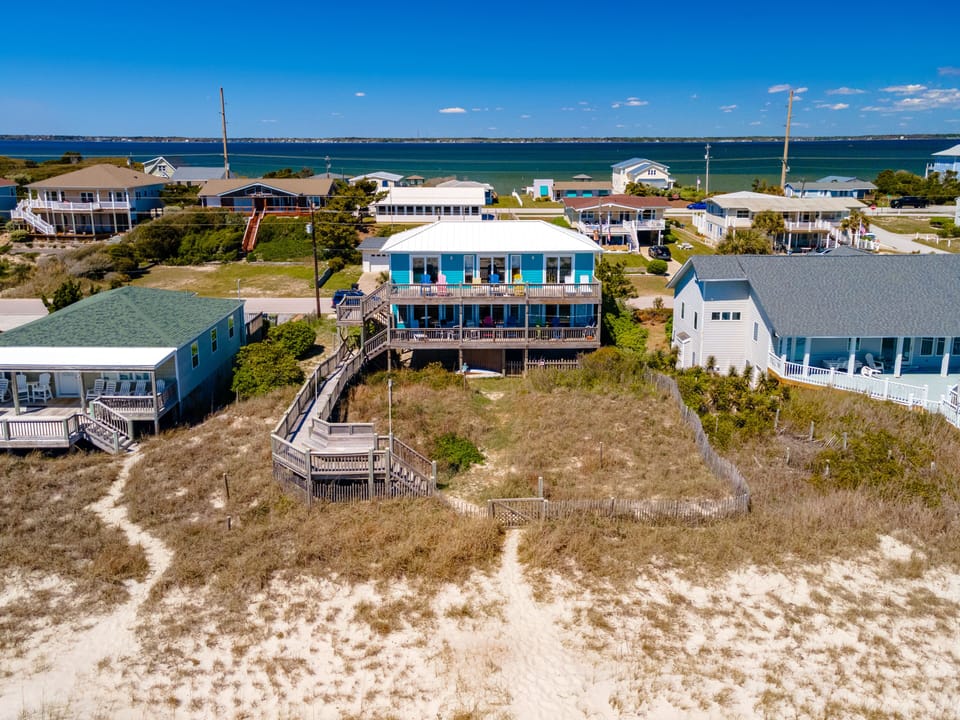 Aerial view of beachfront vacation homes nestled between sandy dunes and sparkling coastal waters in a charming seaside community.