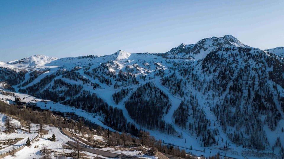 Snow-dusted ski runs carved into a dramatic Alpine mountain under a clear blue sky.