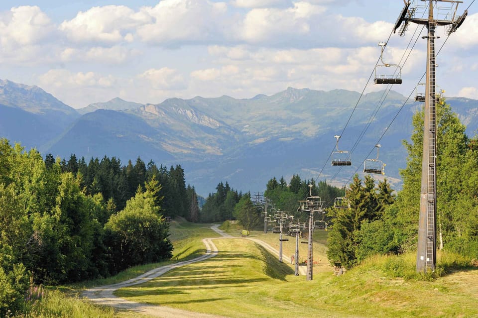 A ski lift trail cutting through lush green Alpine slopes under a bright summer sky.