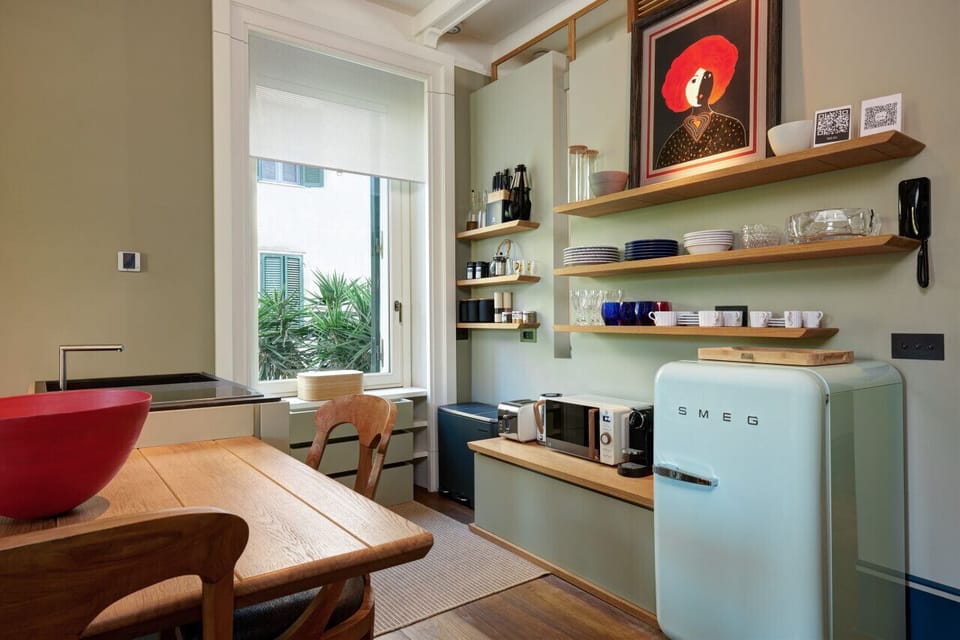 Fully equipped kitchen corner with retro-style Smeg fridge, microwave, coffee machine, and open shelving stocked with essentials. Natural light floods the space, making it ideal for preparing and enjoying homemade meals.