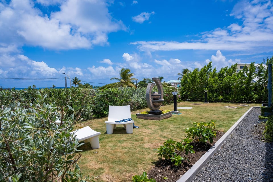 A serene garden featuring a ring-shaped modern sculpture, a white lounge chair, surrounded by lush greenery with an ocean view in the background.